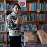 Uczennica klasy VIII prezentuje książkę zapakowaną w szary papier na tle regału z książkami w bibliotece