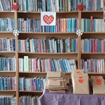 Książki zapakowane w szary papier z naklejonymi czerwonymi serduszkami leżą na stoliku przygotowane do rozpoczęcia akcji na tle regału z książkami w bibliotece