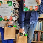 Uczeń i uczennica prezentują dyplomy za konkurs na tle regału z książkami w bibliotece