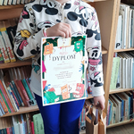 Uczennica klasy prezentuje dyplom za konkurs   na tle regału z książkami w bibliotece